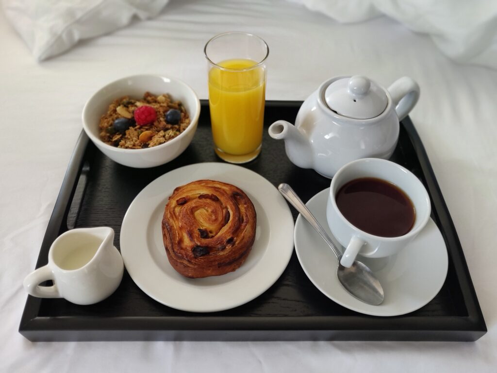 A breakfast tray in bed with orange juice, a pain aux raisins, granola, a teapot and cup of tea
