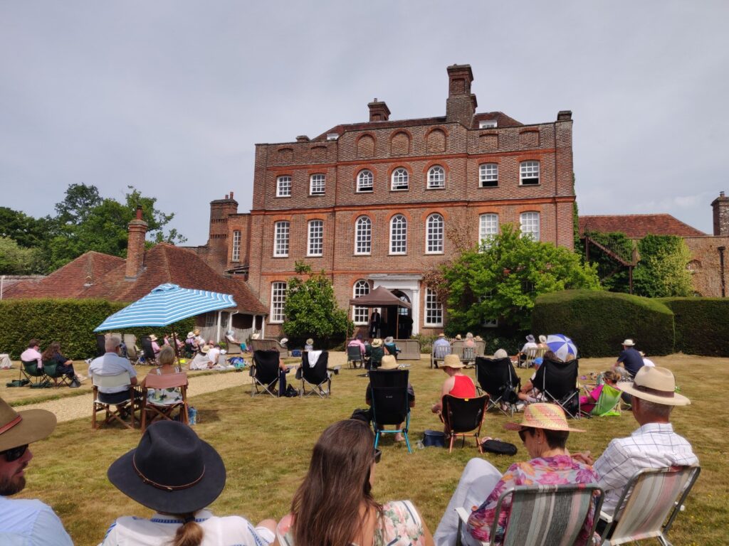 Finchcocks west elevation with Warren Mailley-Smith introducing a piece on the open-air stage. Audience members fill the lawn on deck chairs, picnic blankets and benches.