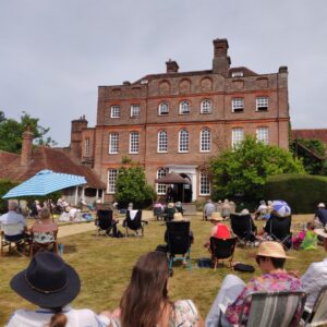 Finchcocks west elevation with Warren Mailley-Smith introducing a piece on the open-air stage. Audience members fill the lawn on deck chairs, picnic blankets and benches.