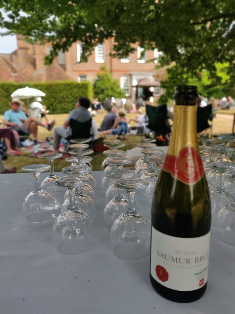 Bottle of champagne in the foreground with wine glasses. Audience and Finchcocks open-air stage in the background.