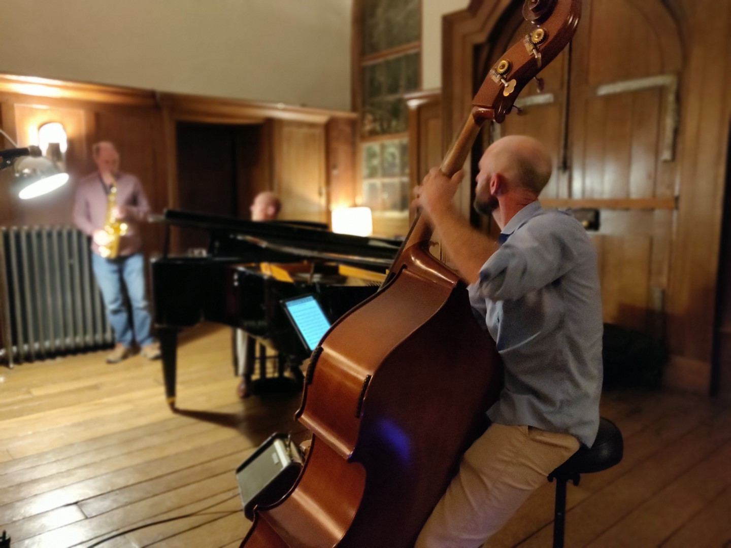 Jazz trio performing in Finchcocks main hall comprising of double bass in the foreground, saxophone and grand piano in the background.