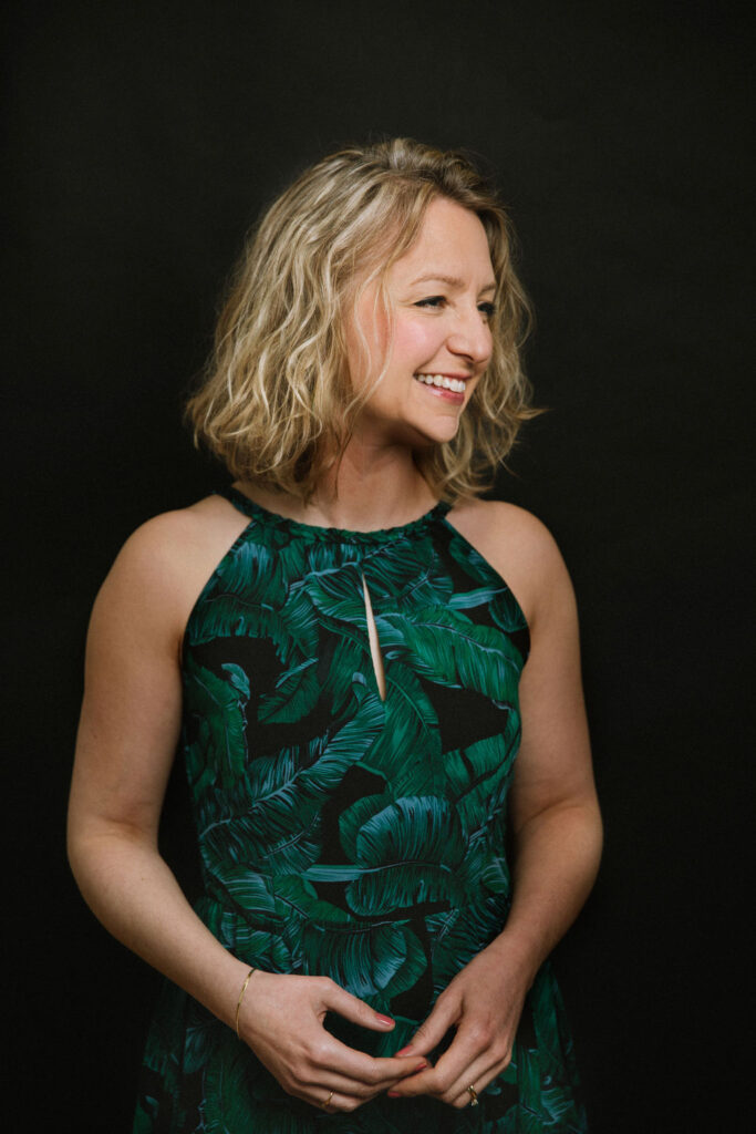 Jennifer Maslin looks to her left smiling. She stands in front of a black background wearing her blonde hair down and a green summer dress.